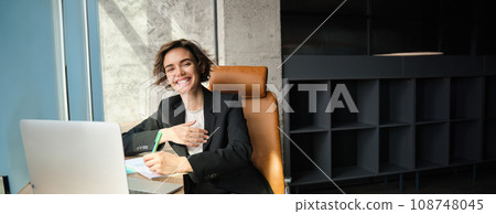 Portrait of woman in an office, working on laptop, making notes, sitting near window 108748045