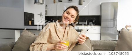 Gorgeous young woman at home, wearing bathrobe, holding mobile phone, using social media app on smartphone, drinking orange juice, sitting on sofa Gorgeous young woman at home, wearing bathrobe, holding mobile phone, using social media app on smartphone, drinking orange juice, sitting on sofa 108748063
