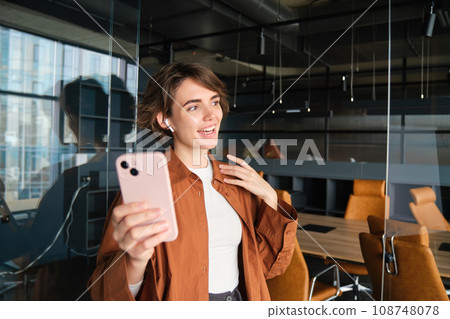 Image of young woman, entrepreneur in office, standing with smartphone, talks online, joins meeting in mobile app, has conversation, wears wireless earphones 108748078