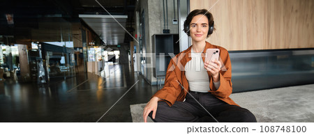 Portrait of female manager, sitting in office on floor, holding smartphone, listening music in wireless headphones 108748100
