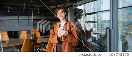 Portrait of smiling beautiful woman, manager in wireless headphones, holding smartphone, working in office 108748150