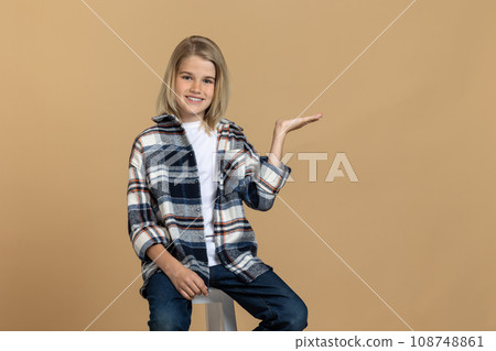 Cute blonde teen girl in checkered shirt sitting on the chair in a studio Cute blonde teen girl in checkered shirt sitting on the chair in a studio 108748861