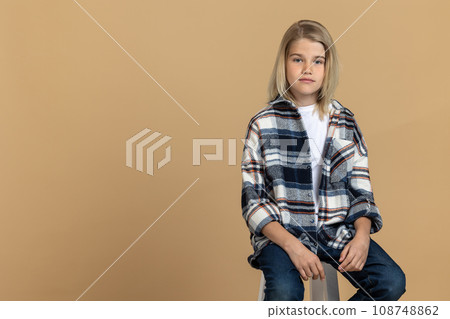 Cute blonde teen girl in checkered shirt sitting on the chair in a studio Cute blonde teen girl in checkered shirt sitting on the chair in a studio 108748862