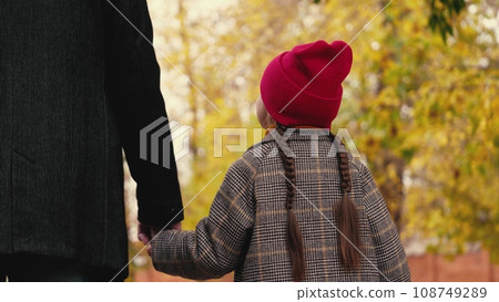 Happy little girl looks at father walking together across autumn garden. Adorable little daughter holds loving father hand walking in park. Father and little daughter rest in garden on autumn day Happy little girl looks at father walking together across autumn garden. Adorable little daughter holds loving father hand walking in park. Father and little daughter rest in garden on autumn day 108749289