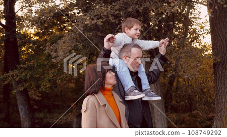 Father carries son on shoulders enjoying serene stroll through park near wife. Father carries son on shoulders while walking alongside wife. Father with son on shoulders enjoys company of wife 108749292