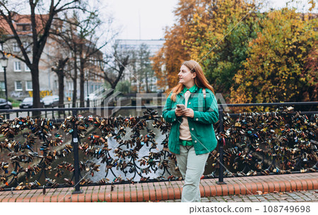 Attractive young female tourist is exploring new city. Gdansk old town, A happy woman is standing near Radunia Canal, travel to Europe. Portrait of beautiful 30s girl using smartphone outdoors. 108749698