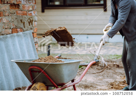 Human in tracksuit and gloves holding spade and throw earth into wheelbarrow. Gardener removing land from lot on yard. 108752058