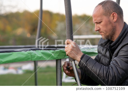 Middle aged human with furrowed eyebrows carefully fastening metal posts to trampoline support outdoor using bolt. Middle aged human with furrowed eyebrows carefully fastening metal posts to trampoline support outdoor using bolt. 108752109