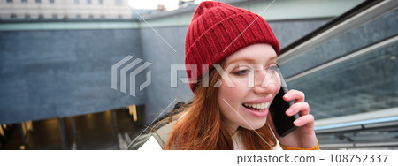 Portrait of young redhead woman walks around city, goes up stairs with mobile phone, talks on smartphone and smiles 108752337