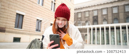 Social media and people. Young redhead girl sits on street, uses mobile phone app, looks up information in internet, holds smartphone Social media and people. Young redhead girl sits on street, uses mobile phone app, looks up information in internet, holds smartphone 108752413