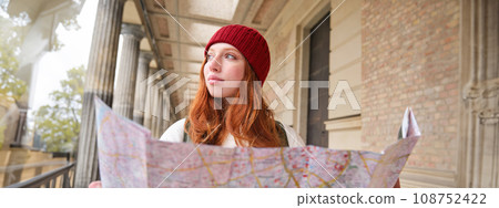 Smiling young redhead woman in red hat, looks at paper map to look for tourist attraction. Tourism and people concept. Girl explores city, tried to find way 108752422