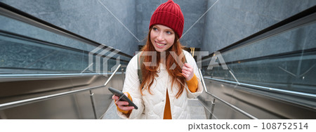 Portrait redhead girl tourist, goes up stairs with smartphone, follows route on mobile phone app, holds backpack and smiles Portrait redhead girl tourist, goes up stairs with smartphone, follows route on mobile phone app, holds backpack and smiles 108752514