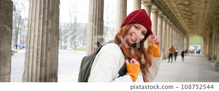 Cute smiling girl walks around city in red hat and gloves, has backpack, tourist explores strees of european city Cute smiling girl walks around city in red hat and gloves, has backpack, tourist explores strees of european city 108752544