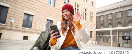 Social media and people. Young redhead girl sits on street, uses mobile phone app, looks up information in internet, holds smartphone 108752545