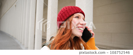 Happy redhead woman, girl with phone talks, has conversation on mobile app, uses internet to call abroad with smartphone app, laughing and smiling Happy redhead woman, girl with phone talks, has conversation on mobile app, uses internet to call abroad with smartphone app, laughing and smiling 108752546
