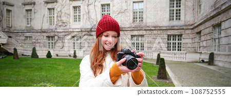 Happy redhead girl tourist, takes photos, photographer with professional camera walks around city and captures beautiful pictures 108752555