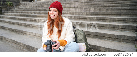 Young student, photographer sits on street stairs and checks her shots on professional camera, taking photos outdoors 108752584