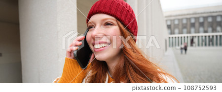 Happy redhead woman, girl with phone talks, has conversation on mobile app, uses internet to call abroad with smartphone app, laughing and smiling 108752593