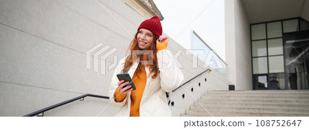 Happy girl student in red hat, holds smartphone, tourist looks at map app on her phone, explores sightseeing, texts message, looks for couchsurfing, rents place to stay online Happy girl student in red hat, holds smartphone, tourist looks at map app on her phone, explores sightseeing, texts message, looks for couchsurfing, rents place to stay online 108752647