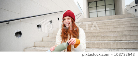 Smiling traveler, redhead girl tourist sits on stairs with flask, drinks hot coffee from thermos while travelling and sightseeing around foreign city, sits on stairs and rests 108752648