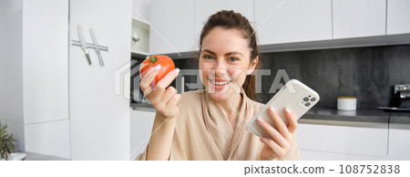 Image of smiling beautiful woman, sits in kitchen, holds smartphone and tomato, looks happy, orders groceries delivery to her home, using mobile phone app, buying online vegetables for her recipe Image of smiling beautiful woman, sits in kitchen, holds smartphone and tomato, looks happy, orders groceries delivery to her home, using mobile phone app, buying online vegetables for her recipe 108752838