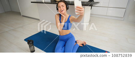 Beautiful and stylish sportswoman, posing for selfie, listens music in wireless headphones, holds smartphone, sits on fitness mat, workout from home 108752839