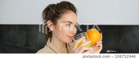 young brunette woman drinking orange juice in white modern kitchen and smiling, girl holding glass of fresh juice, healthy food and drink concept 108752946