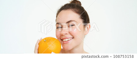 Close up portrait of smiling young woman, holding orange near face, looking happy, concept of vitamins, organic and natural skincare treatment with C serum, white background Close up portrait of smiling young woman, holding orange near face, looking happy, concept of vitamins, organic and natural skincare treatment with C serum, white background 108753158