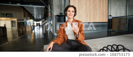 Portrait of happy young woman, manager in office, sitting in lounge room, relaxing on lunch in break room, holding smartphone, listening music in wireless headphones, using co-working space Portrait of happy young woman, manager in office, sitting in lounge room, relaxing on lunch in break room, holding smartphone, listening music in wireless headphones, using co-working space 108753173