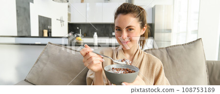 Close up portrait of cute woman on sofa, eating breakfast, holding delicious chocolate cereals with milk in a bowl, having proper morning meal Close up portrait of cute woman on sofa, eating breakfast, holding delicious chocolate cereals with milk in a bowl, having proper morning meal 108753189