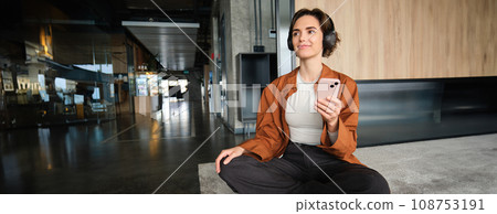 Image of stylish young company employee, sitting in office on floor, resting in lounge room at co-working space, holding mobile phone, wearing headphones 108753191