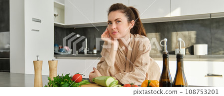 Portrait of pretty woman in bathrobe, standing in kitchen with thoughtful face, posing near vegetables and bottle of oil, thinking what food to prepare, making salad, eating healthy 108753201