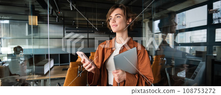 Portrait of office woman, working digital nomad standing in an office, using smartphone, holding laptop Portrait of office woman, working digital nomad standing in an office, using smartphone, holding laptop 108753272