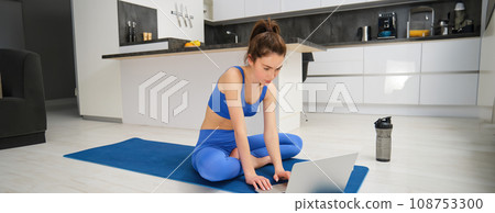 Image of young fitness instructor using laptop for online workout training, showing fitness exercises, talking to client, sitting on yoga rubber mat in sportswear 108753300