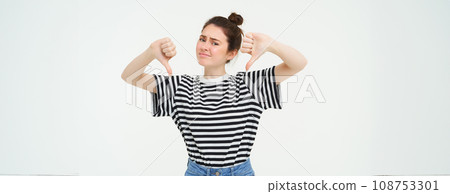 Young woman gives negative feedback, shows thumbs down, disapproves product, dislikes smth bad, stands over white background Young woman gives negative feedback, shows thumbs down, disapproves product, dislikes smth bad, stands over white background 108753301