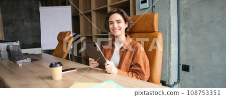 Portrait of young woman CEO, businesswoman in casual clothes, sitting in her office with digital tablet and cup of coffee, working on project, reading Portrait of young woman CEO, businesswoman in casual clothes, sitting in her office with digital tablet and cup of coffee, working on project, reading 108753351