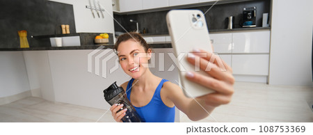 Wellbeing and sports. Beautiful young woman taking selfie on smartphone, doing fitness training from home, drink water, stays hydrated and doing exercises 108753369