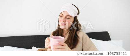 Gorgeous woman in pyjamas and sleeping mask, holding cup of coffee, drinking cappuccino in her bed and looking happy, resting in bedroom on weekend, having relaxing morning Gorgeous woman in pyjamas and sleeping mask, holding cup of coffee, drinking cappuccino in her bed and looking happy, resting in bedroom on weekend, having relaxing morning 108753411