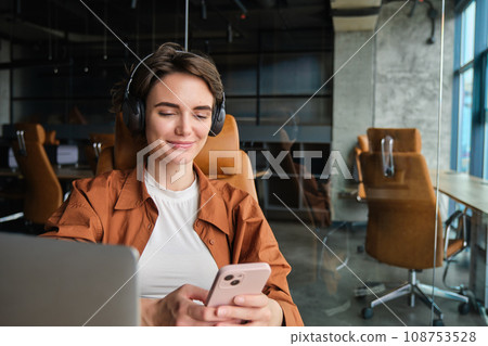 Portrait of female employee, entrepreneur in office with laptop, mobile phone, wears wireless headphones Portrait of female employee, entrepreneur in office with laptop, mobile phone, wears wireless headphones 108753528
