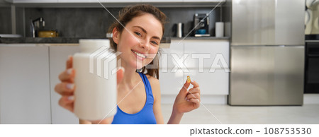 Close up portrait of fitness woman, girl workout from home, showing bottle of vitamins, dietary supplement pills, recommending buds Close up portrait of fitness woman, girl workout from home, showing bottle of vitamins, dietary supplement pills, recommending buds 108753530