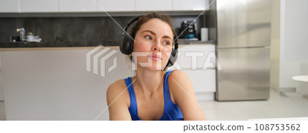 Beautiful brunette girl, doing fitness workout, sitting in living room in wireless headphones, listening to music and doing sports Beautiful brunette girl, doing fitness workout, sitting in living room in wireless headphones, listening to music and doing sports 108753562