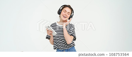 Young woman with smartphone listening to music, dancing to her favourite song in headphones, posing against white background Young woman with smartphone listening to music, dancing to her favourite song in headphones, posing against white background 108753564