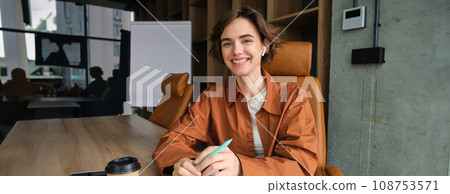 Portrait of young cheerful woman working on start up project, making notes, holding pen and planner, reading documents, sitting in an office 108753571