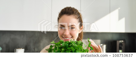 Portrait of beautiful smiling girl with bouquet of parsley, standing in the kitchen and cooking, adding herbs to healthy fresh salad or meal, preparing food 108753637