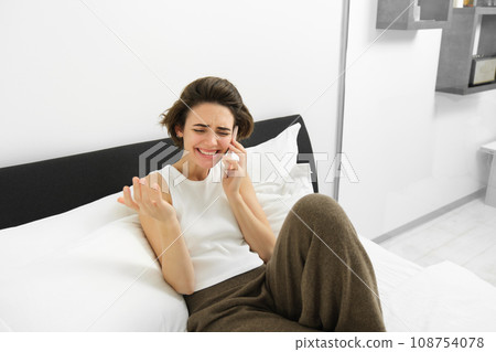 Carefree young woman lying in bed, talking on mobile phone, calling friend, chatting while resting in bedroom, laughing and smiling during conversation 108754078