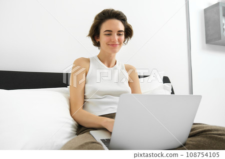 Portrait of smiling modern woman, sitting on her bed with laptop, looking happy, working from home, checking her emails on computer, browsing online shops Portrait of smiling modern woman, sitting on her bed with laptop, looking happy, working from home, checking her emails on computer, browsing online shops 108754105