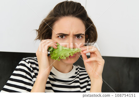 Frowning silly woman, holding lettuce leaf near face and frowning at camera, making pouting face, dislike eating vegetables 108754151