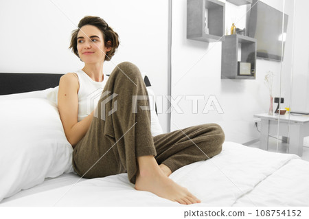 Portrait of happy, relaxed young woman resting in hotel room, lying in bed in her cosy pyjamas, looking around 108754152