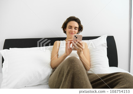 Portrait of young smiling woman resting in her bedroom, lying in bed and using smartphone, online shopping on mobile phone, scrolling social media on device 108754153
