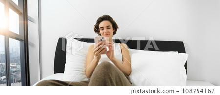 Portrait of young smiling woman resting in her bedroom, lying in bed and using smartphone, online shopping on mobile phone, scrolling social media on device Portrait of young smiling woman resting in her bedroom, lying in bed and using smartphone, online shopping on mobile phone, scrolling social media on device 108754162